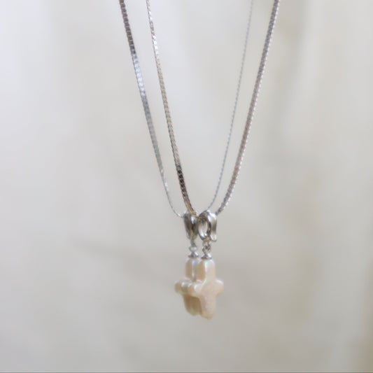 A silver-toned chain necklace with a small cream-colored cross pendant, displayed against a soft, light beige background.