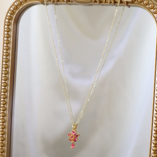A delicate gold chain necklace with a small pink floral pendant, hanging in front of an ornate gold mirror.