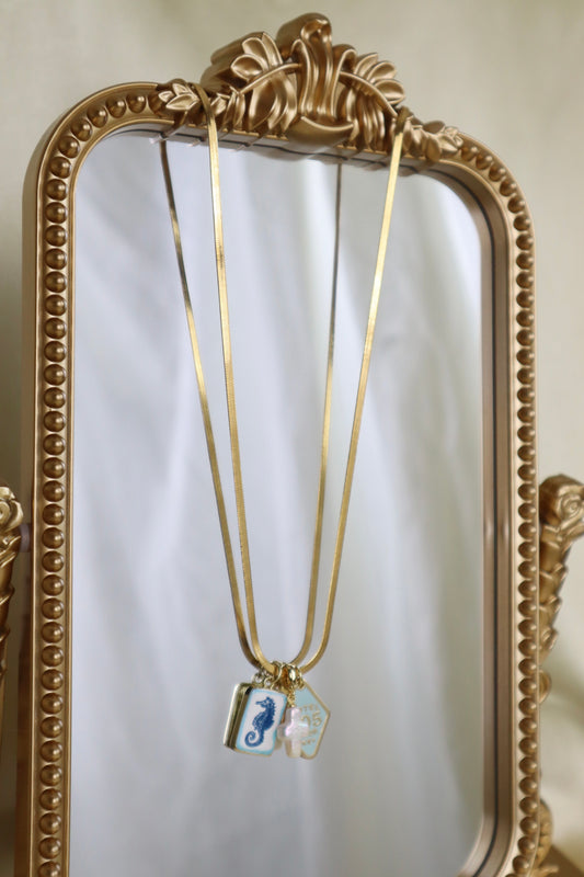 Gold necklace with two small pendants, one featuring a blue seahorse and the other a white seahorse, hanging in front of an ornate gold mirror.