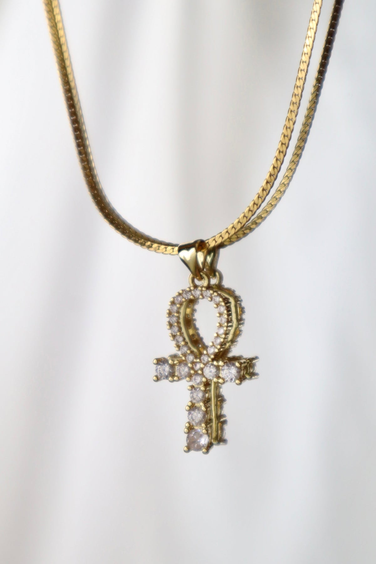 A gold-tone chain necklace featuring an Ankh pendant adorned with small clear stones, displayed against a soft white draped background.