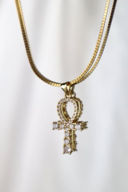A gold-tone chain necklace featuring an Ankh pendant adorned with small clear stones, displayed against a soft white draped background.