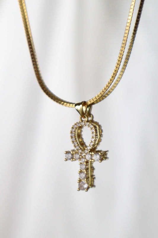 A gold-tone chain necklace featuring an Ankh pendant adorned with small clear stones, displayed against a soft white draped background.