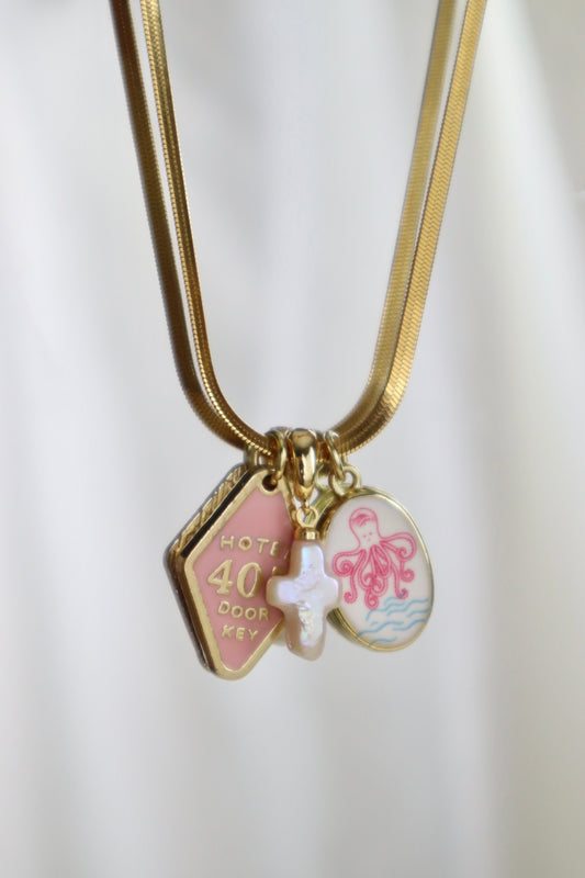 Gold chain necklace with three charms: a pink house-shaped charm reading 'HOTEL 40 COOK KEY,' a white cross charm, and an octopus-themed round charm on a white background.