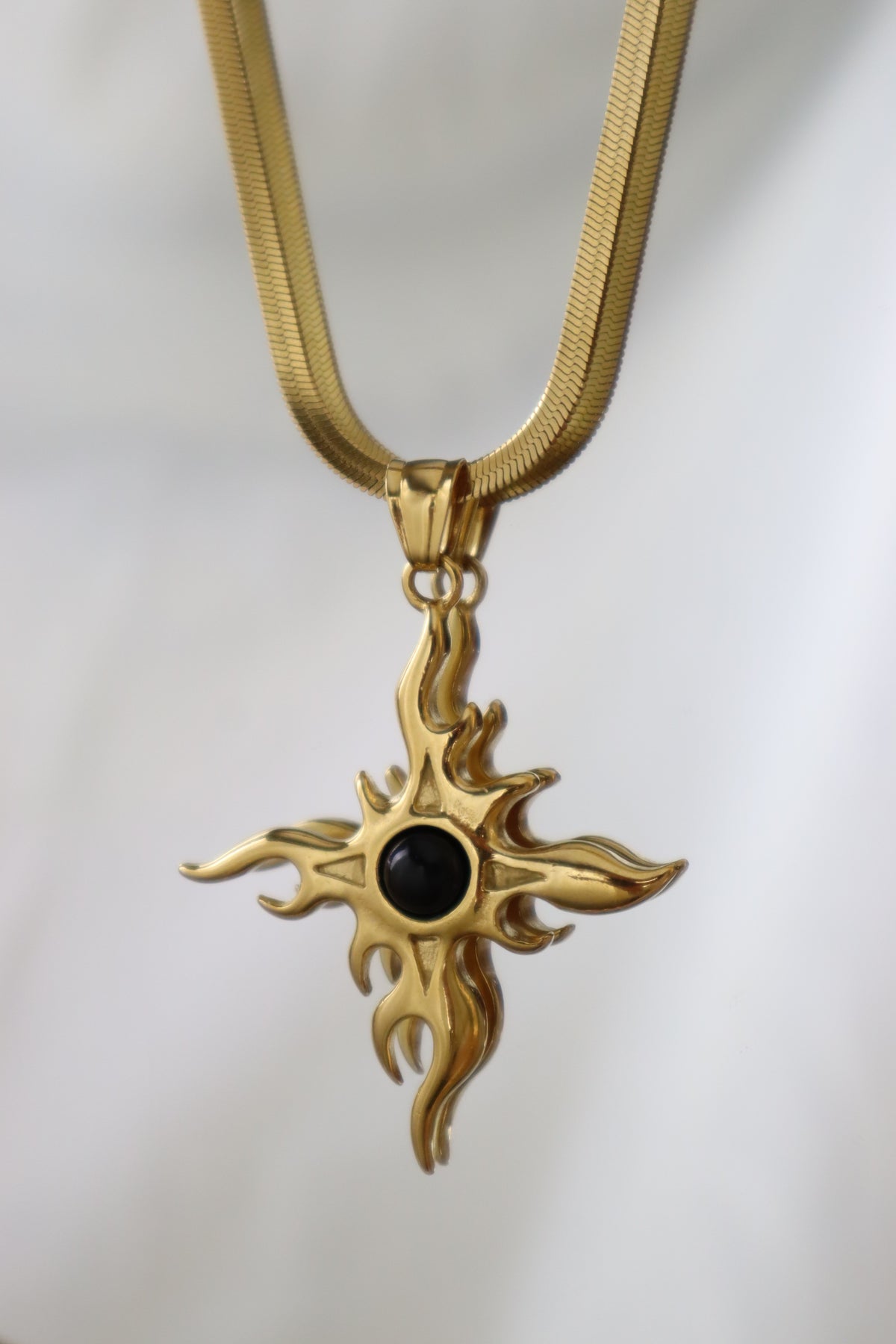 A gold-toned cross pendant necklace with a central black oval stone, suspended from a matching gold chain against a soft white background.