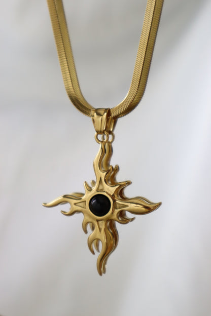 A gold-toned cross pendant necklace with a central black oval stone, suspended from a matching gold chain against a soft white background.