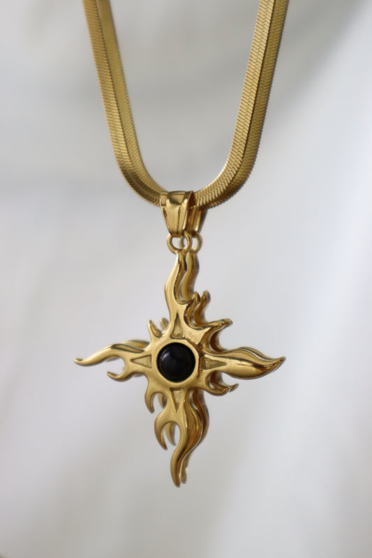 A gold-toned cross pendant necklace with a central black oval stone, suspended from a matching gold chain against a soft white background.