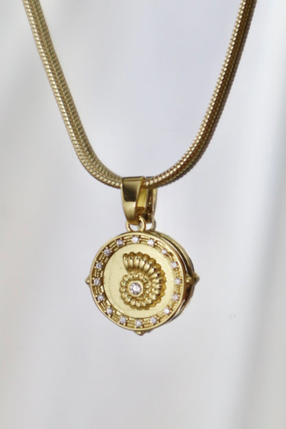 A gold necklace with a round pendant featuring a spiral shell design and a central clear gemstone, set against a plain light background.