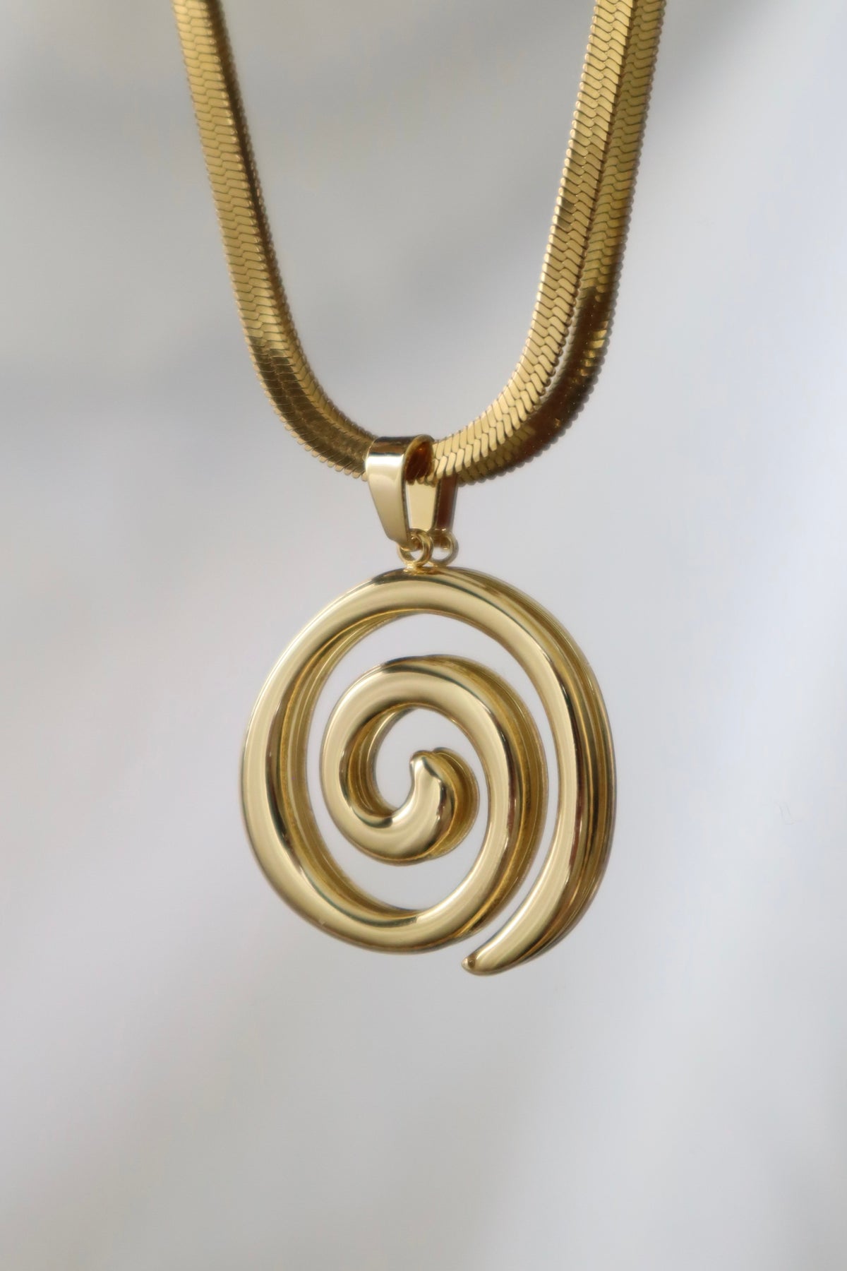 A gold spiral pendant necklace on a textured gold snake chain, shown against a soft, light gray background.