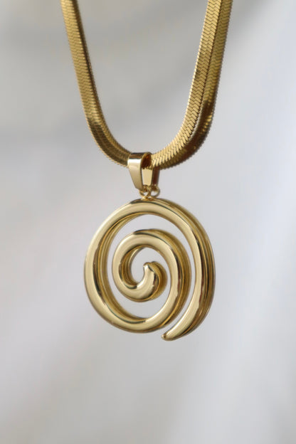 A gold spiral pendant necklace on a textured gold snake chain, shown against a soft, light gray background.