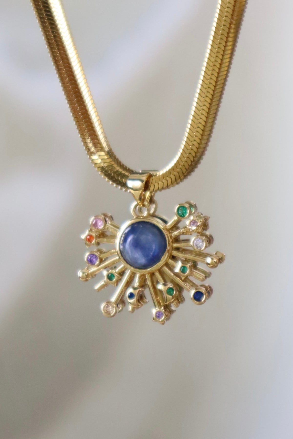 A gold snake chain necklace with a sunburst pendant featuring a central blue oval stone and small multicolored gemstone accents, shown against a soft light gray background.