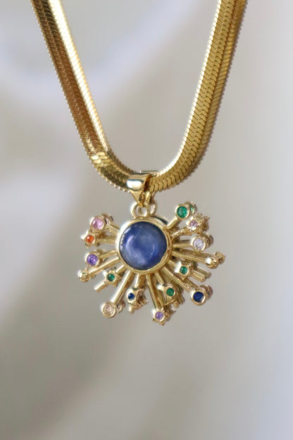 A gold snake chain necklace with a sunburst pendant featuring a central blue oval stone and small multicolored gemstone accents, shown against a soft light gray background.