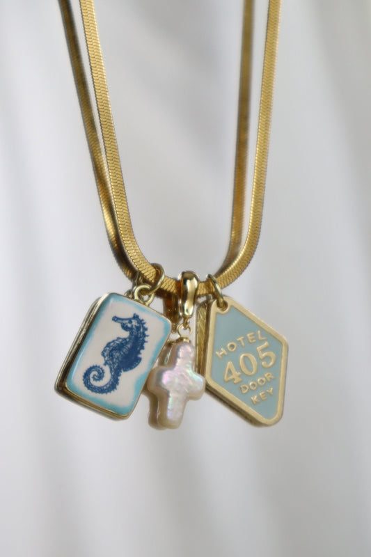 A gold chain necklace with three charms: a light blue rectangular charm with a dark blue seahorse, a light blue shield-shaped charm with gold text reading 'HOTEL 405', and a small iridescent pink cross charm, shown against a white draped fabric background.