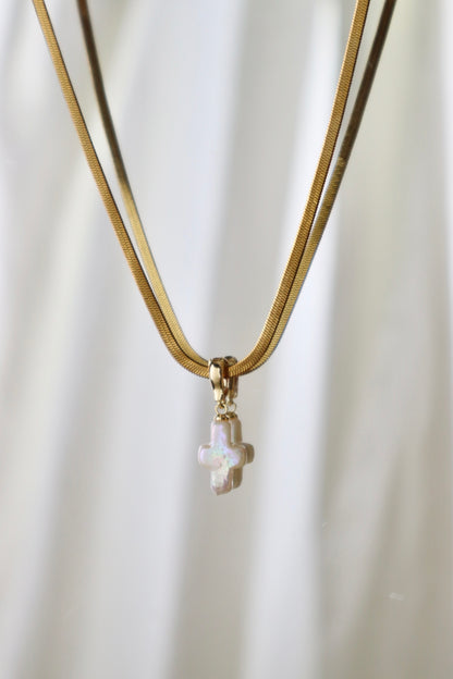 A gold snake chain necklace featuring a small gold cross pendant with a white iridescent opal inlay, set against a soft white draped fabric background.