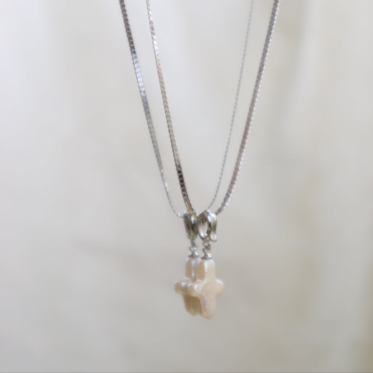 A silver-toned chain necklace with a small cream-colored cross pendant, displayed against a soft, light beige background.