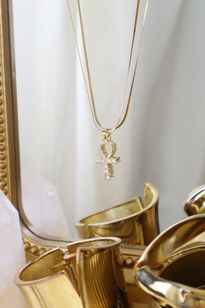 A gold chain necklace with an ankh pendant adorned with small clear stones, displayed in front of a gold mirror with reflective gold accessories.