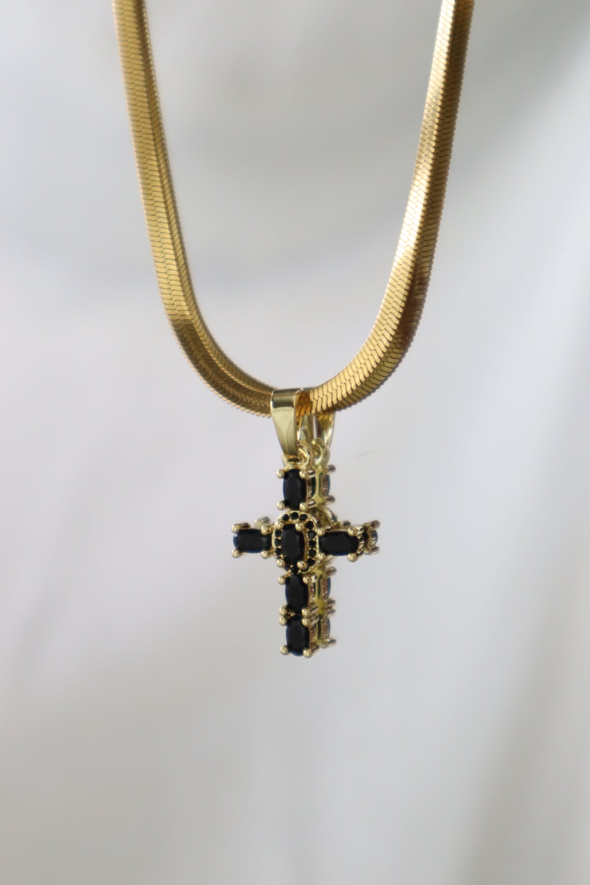 A gold snake chain necklace with a gold cross pendant featuring black rectangular stones, shown against a plain light gray background.