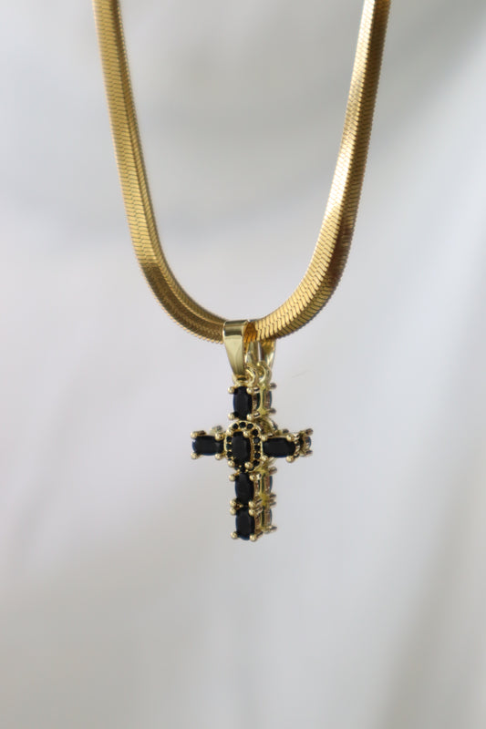A gold snake chain necklace with a gold cross pendant featuring black rectangular stones, shown against a plain light gray background.