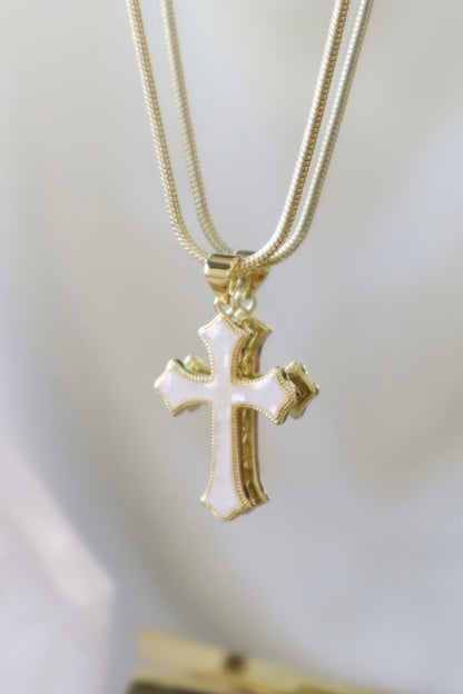 Gold cross pendant necklace with white enamel detailing, hanging on a gold snake chain against a soft white background.