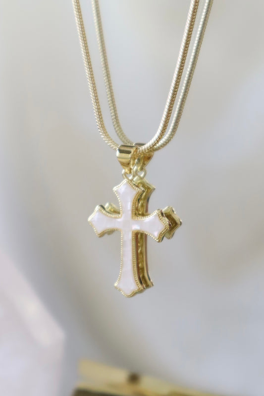 Gold cross pendant necklace with white enamel detailing, hanging on a gold snake chain against a soft white background.