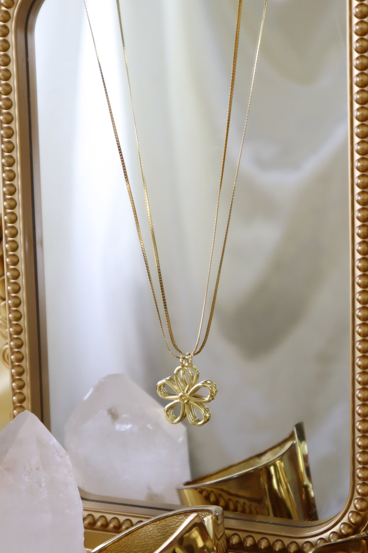 A gold-tone chain necklace with a four-petal flower pendant, displayed in front of a decorative gold mirror with a white satin backdrop.