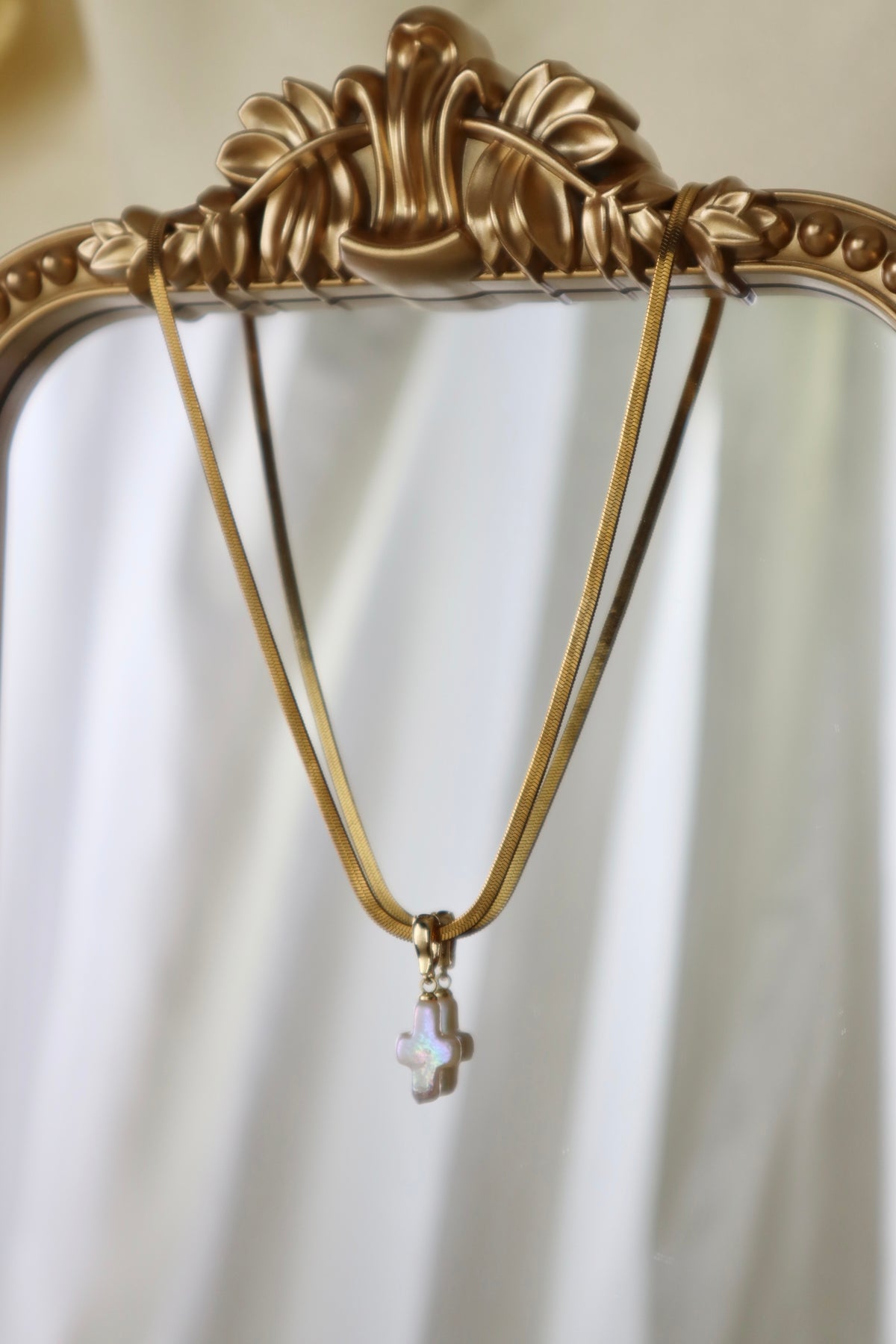 A gold-tone necklace with a small iridescent cross pendant hanging from an ornate gold mirror frame against a soft white draped background.