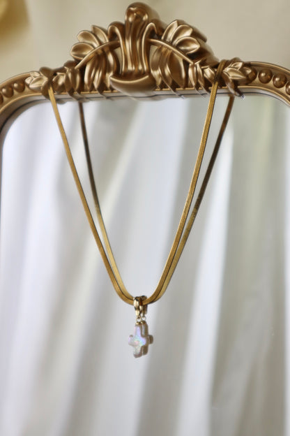 A gold-tone necklace with a small iridescent cross pendant hanging from an ornate gold mirror frame against a soft white draped background.