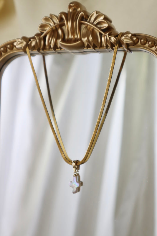 A gold-tone necklace with a small iridescent cross pendant hanging from an ornate gold mirror frame against a soft white draped background.