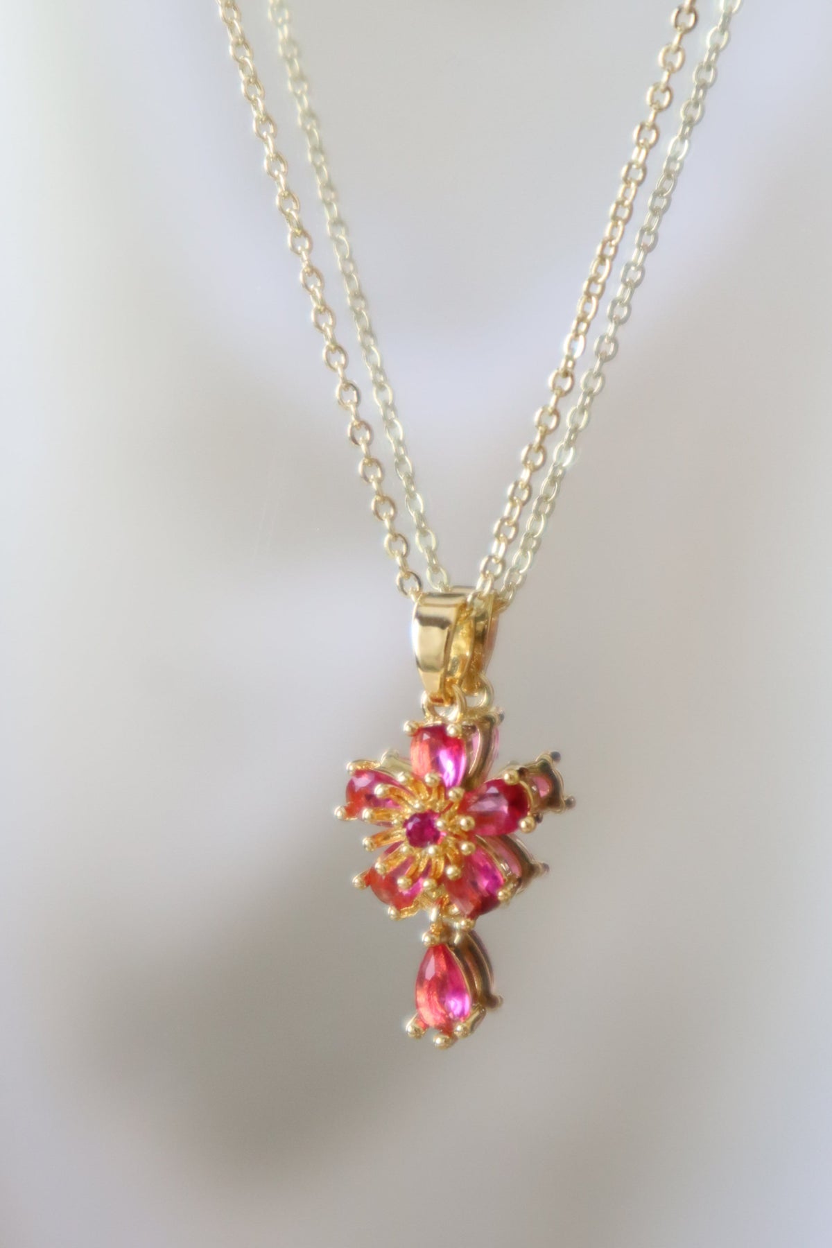 A gold cross pendant necklace adorned with sparkling pink gemstones, displayed against a soft, out-of-focus white background.