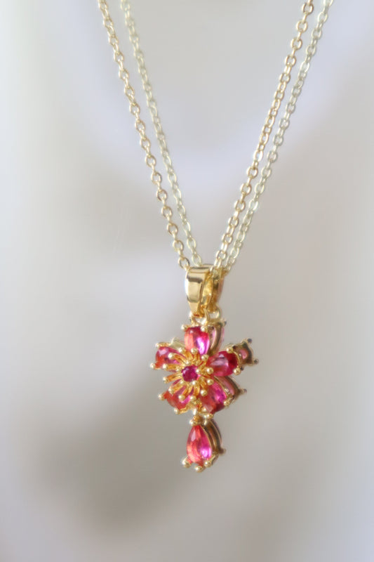 A gold cross pendant necklace adorned with sparkling pink gemstones, displayed against a soft, out-of-focus white background.