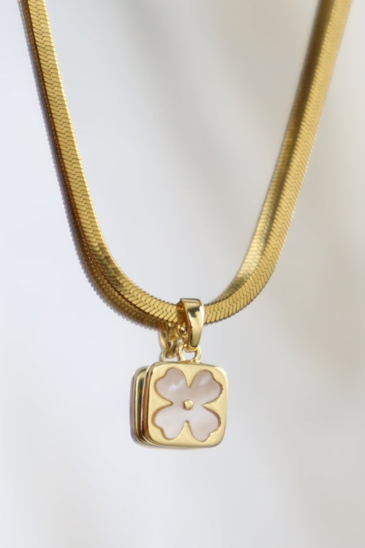 A gold snake chain necklace with a square pendant featuring a white mother-of-pearl flower cutout, displayed against a soft white background.