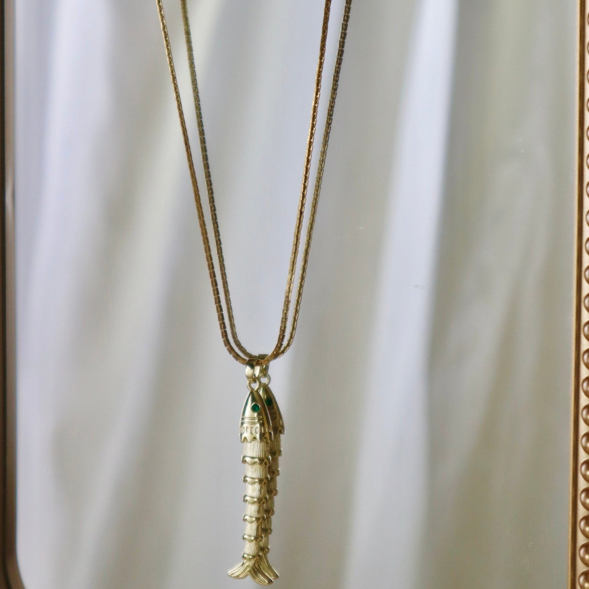A gold-toned fish pendant necklace hanging from a matching chain, displayed against a reflective gold-framed mirror with a white draped backdrop.