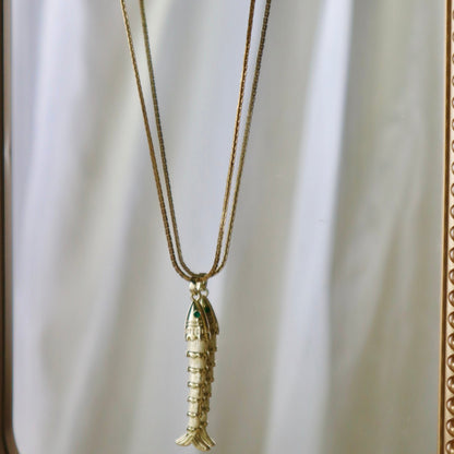 A gold-toned fish pendant necklace hanging from a matching chain, displayed against a reflective gold-framed mirror with a white draped backdrop.