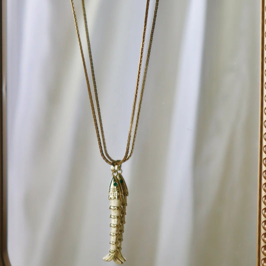 A gold-toned fish pendant necklace hanging from a matching chain, displayed against a reflective gold-framed mirror with a white draped backdrop.