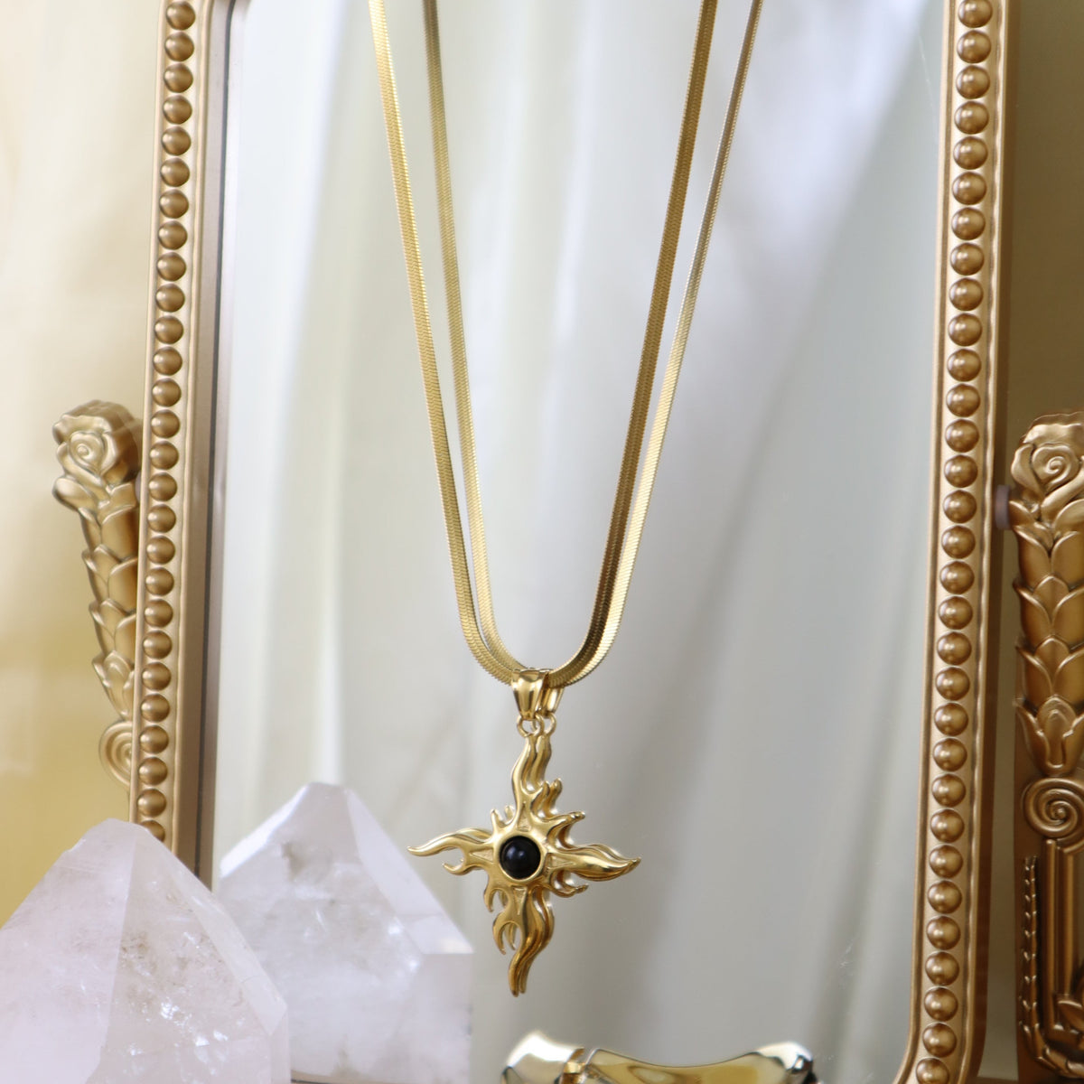 A gold-toned necklace with a decorative cross pendant featuring a small black stone, hanging in front of an ornate gold mirror.