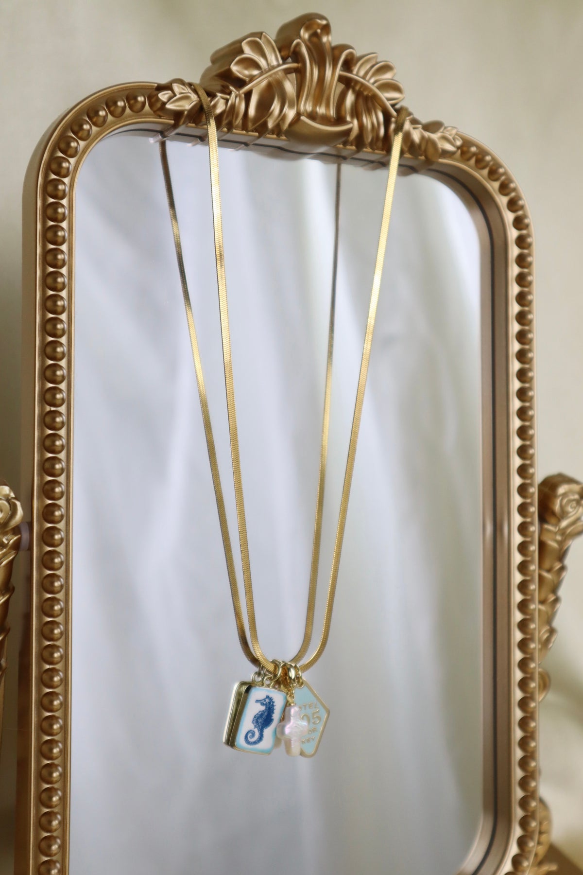 Gold necklace with two small pendants, one featuring a blue seahorse and the other a white seahorse, hanging in front of an ornate gold mirror.