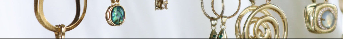 A gold jewelry organizer rack with multiple necklaces hanging from its ornate frame, alongside stacked gold jewelry pieces including bracelets and rings. Green leaves from a plant drape over the top of the rack, and a white background is visible behind it.