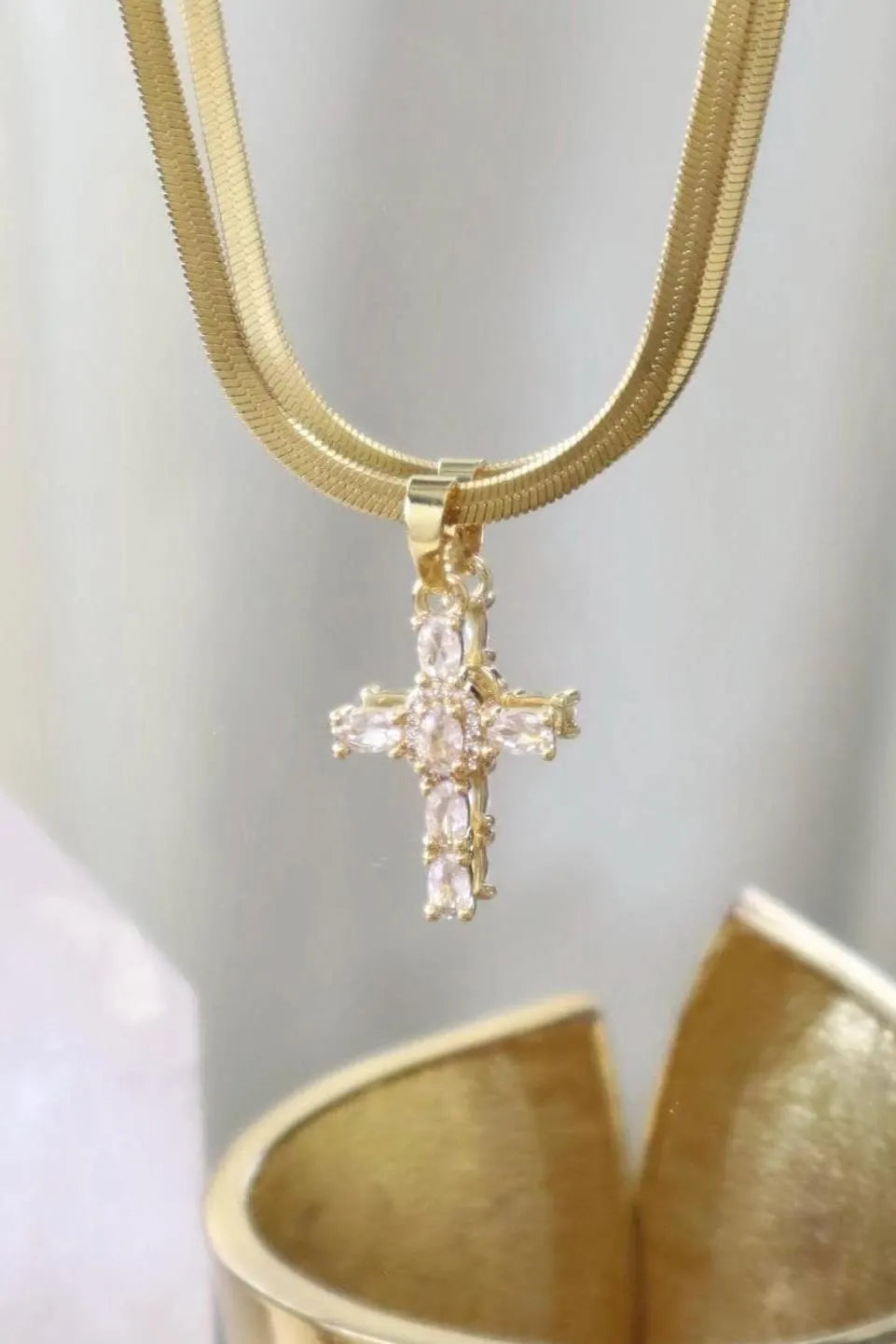 A gold chain necklace with a gold cross pendant adorned with clear rectangular crystals, shown against a soft light gray background with a gold bowl at the bottom.